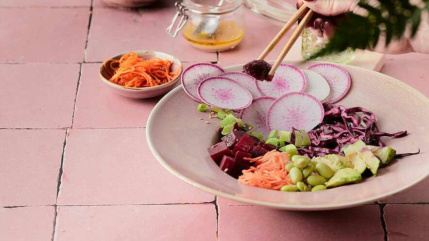 Eine Schüssel mit buntem Salat, gefüllt mit verschiedenen Gemüsesorten wie Radieschen, roten und grünen Bohnen, Karotten und Rotkohl. Eine Hand hält Essstäbchen und nimmt etwas von dem Salat. Im Hintergrund sind weitere Zutaten und ein Glas mit Dressing sichtbar.