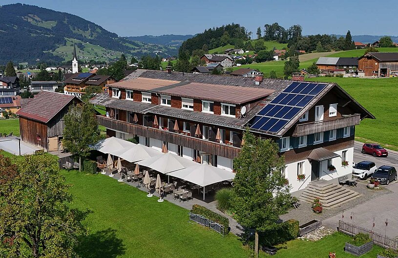 Ein zweigeschossiges, traditionelles Holzgebäude mit einem modernen Dach voller Solarpaneele. Vor dem Haus befinden sich Terrassen mit Sonnenschirmen. Im Hintergrund erstrecken sich grüne Wiesen und sanfte Hügel. Die Umgebung strahlt eine ruhige, ländliche Atmosphäre aus.
