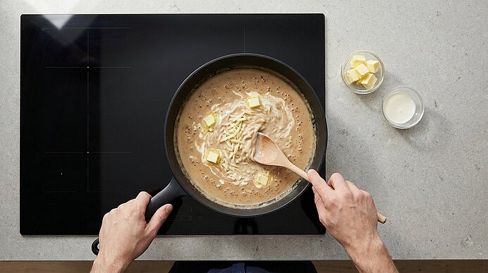 Draufsicht: Hände rühren mit einem Holzlöffel in einer Pfanne eine cremige Pasta-Soße mit Butterwürfeln und geriebenem Käse auf einem Kochfeld.