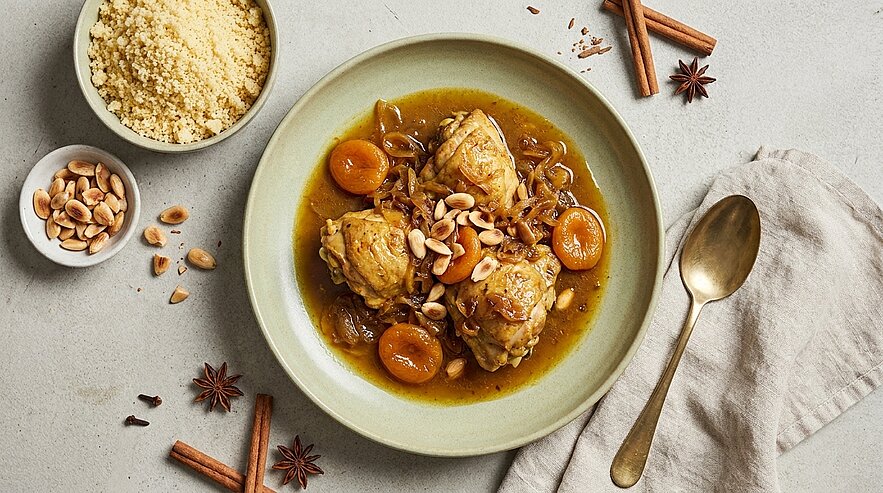 Teller mit marokkanisch gewürzten Hähnchenstücken in aromatischer Sauce, getrockneten Aprikosen, karamellisierten Zwiebeln und Erdnüssen. Daneben eine Schale Couscous.