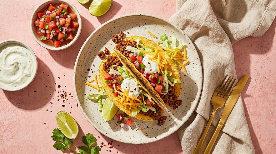Zwei knusprige Tacos mit gewürztem Hackfleisch, Salat, geriebenem Käse, Tomaten‑Salsa und Sauerrahm auf einem Teller, dazu Limettenstücke.