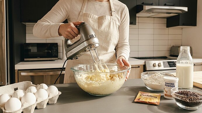 Person rührt mit elektrischem Handmixer Teig in einer Glasschüssel; auf der Arbeitsplatte liegen Eier, Milch, Mehl, Backpulver und Schokostreusel.