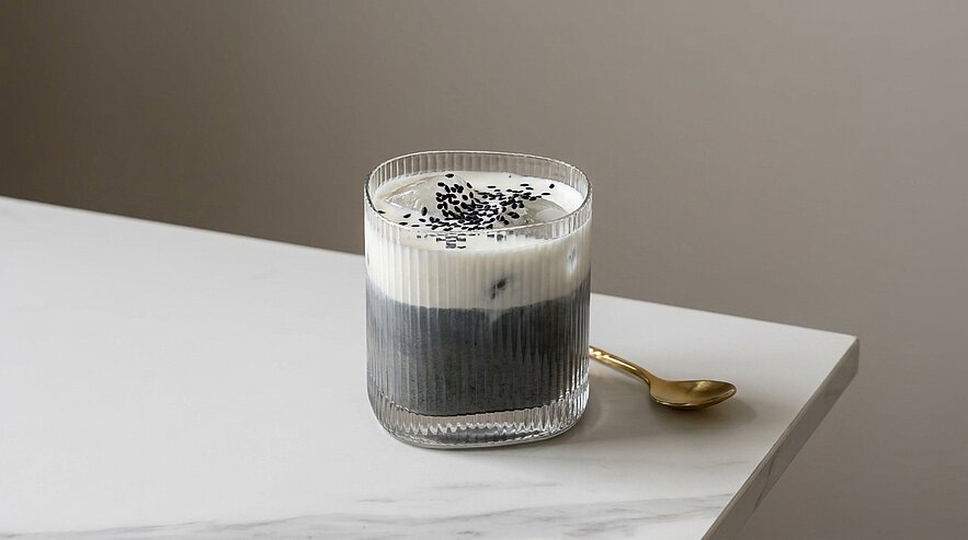 Glas mit zweischichtiger Creme: dunkle, fast schwarze Unterlage und weiße Oberschicht, mit schwarzen Sesamsamen bestreut; ein goldener Löffel liegt daneben auf dem Tisch.