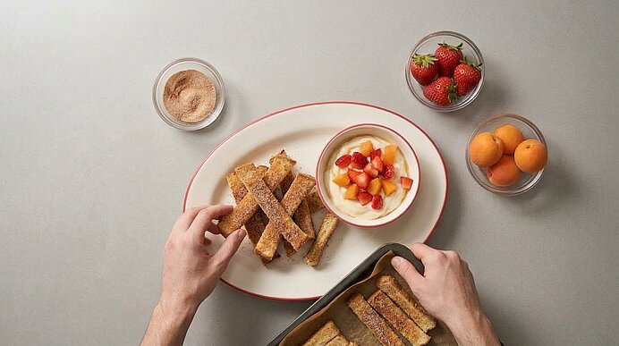 Zwei Hände arrangieren gebackene Toaststifte auf einer Servierplatte neben einer Schale Fruchtquark.