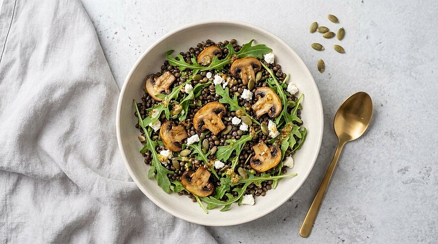 Schüssel mit Linsensalat aus Belugalinsen, Rucola, gebratenen Champignons, Feta und Kürbiskernen.