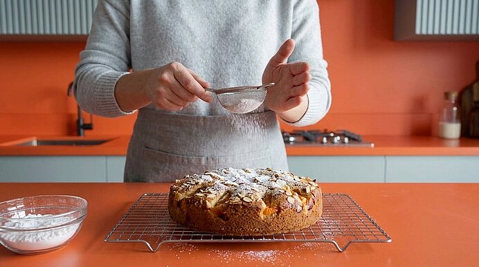 Hände einer Person streuen Puderzucker über einen runden, frisch gebackenen Kuchen auf einem Abkühlgitter in einer Küche.