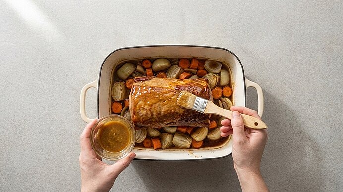 Zwei Hände bestreichen einen gebratenen Braten in einer Auflaufform mit Glasur; ein Backpinsel und eine Schüssel mit Sauce sind zu sehen. Um den Braten liegen Karotten und halbierte Zwiebeln.