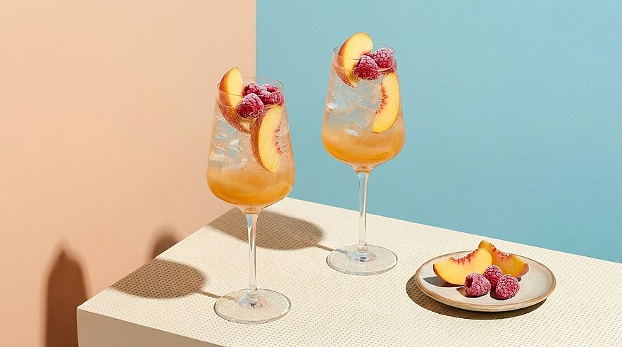 Zwei Gläser mit einem fruchtigen Getränk stehen auf einem hellen Tisch. Die Gläser sind mit Eis, Pfirsichscheiben und Himbeeren dekoriert. Neben den Gläsern befindet sich ein kleiner Teller mit weiteren Pfirsichscheiben und Himbeeren. Der Hintergrund ist in sanften Farbtönen gehalten.