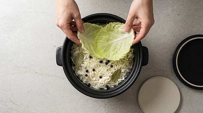 Zwei Hände legen ein Kohlblatt in einen Topf mit geschnittenem Weißkohl, Lorbeerblättern und Pfefferkörnern – Zubereitung von Sauerkraut.