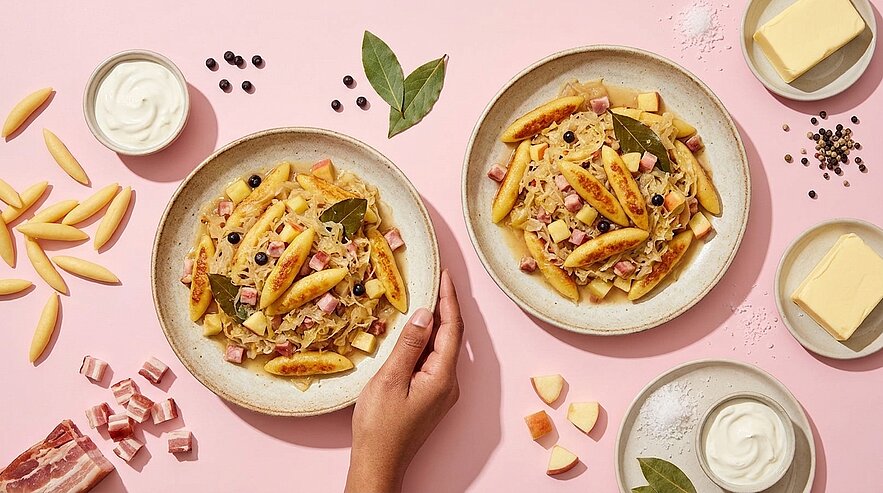 Zwei Schalen Pasta in cremiger Sauce mit gebräunten Nudeln, gewürfeltem Speck, schwarzen Oliven und Lorbeerblatt; eine Hand greift nach einer Schale.