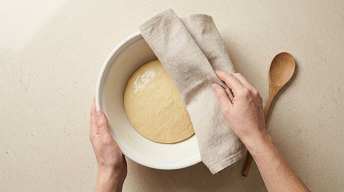 Hände bedecken eine Schüssel mit aufgegangenem Teig mit einem Leinentuch; daneben liegt ein Holzlöffel.