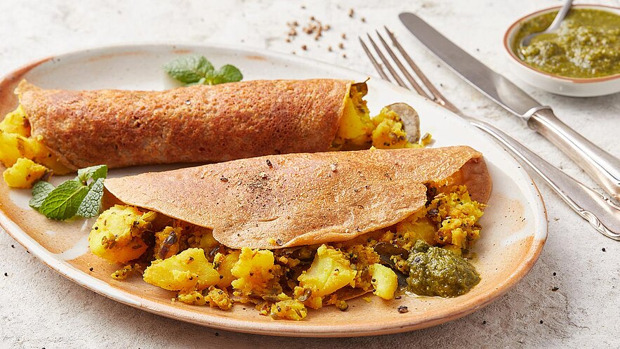 Auf dem Bild sind zwei gefüllte Dosas zu sehen, die auf einem Teller serviert werden. Die Dosas sind aus braunem Teig und sind mit gewürzten Kartoffeln gefüllt. Neben den Dosas liegt etwas grüne Soße. Frische Minzblätter dekorieren den Teller.