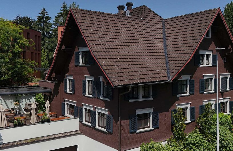 Ein braunes Haus mit einem steilen, roten Dach. Die Fassade hat mehrere Fenster mit weißen Rahmen und blauen Fensterläden. Im Vordergrund gibt es eine Terrasse mit Tischen, Stühlen und Sonnenschirmen. Um das Haus sind Bäume und Sträucher zu sehen, bei klarem, blauem Himmel.