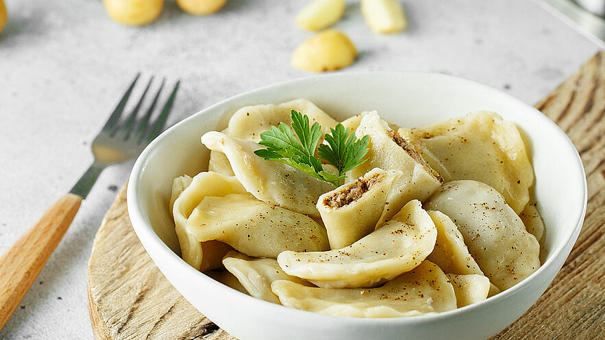 Auf dem Bild ist ein weißer Teller zu sehen, auf dem köstliche Schlutzkrapfen mit Sauerkraut und Pilzen serviert werden. Das Gericht ist mit frischen Spinatblättern dekoriert. Ein Messer liegt am Rand des Tellers. Die Schlutzkrapfen, gefüllt mit herzhaften Zutaten, strahlen eine goldene Farbe aus und verströmen einen verlockenden Duft. 