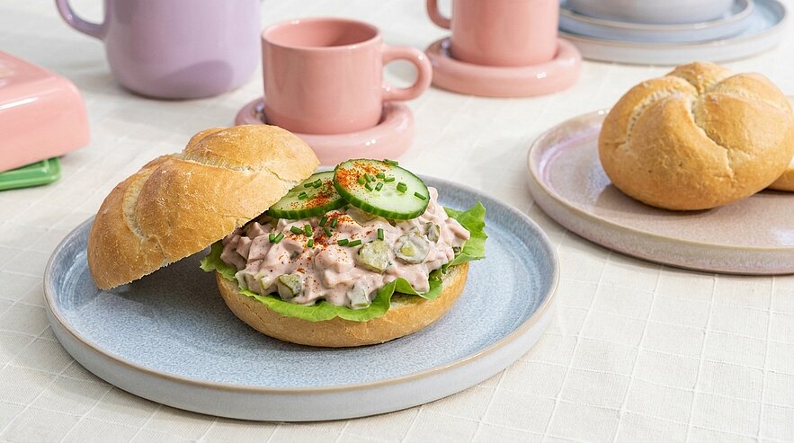 Aufgeschnittenes Brötchen gefüllt mit cremigem Salataufstrich auf einem Salatblatt, garniert mit Gurkenscheiben und Paprikapulver.