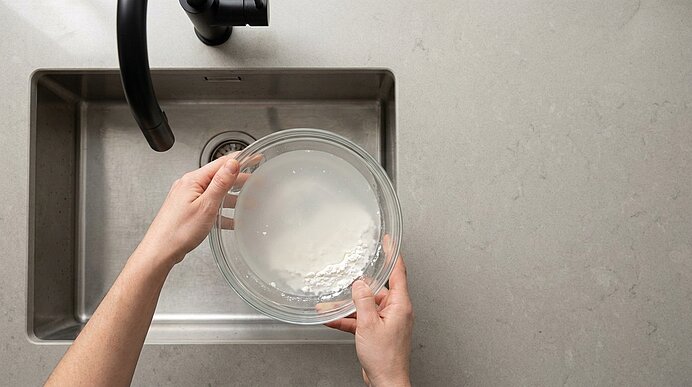 Zwei Hände halten eine Glasschüssel mit trübem, milchigem Wasser über einer Küchenspüle.