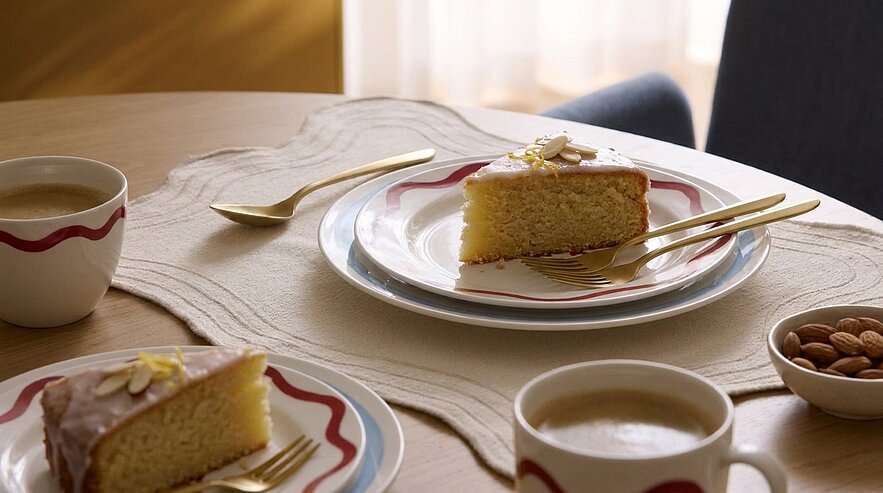Auf dem Tisch liegen zwei Tassen Kaffee und zwei Teller mit einer Scheibe Kuchen, dekoriert mit einer Zuckerglasur und Mandeln. Goldene Bestecke sind auf den Tellern platziert. Der Tisch hat eine textile Unterlage, und im Hintergrund sind weiche Lichtverhältnisse sichtbar.