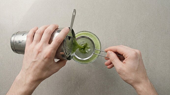 Zwei Hände gießen den grünen Matcha-Cocktail aus einem Shaker durch ein Sieb in ein Coupé-Glas.