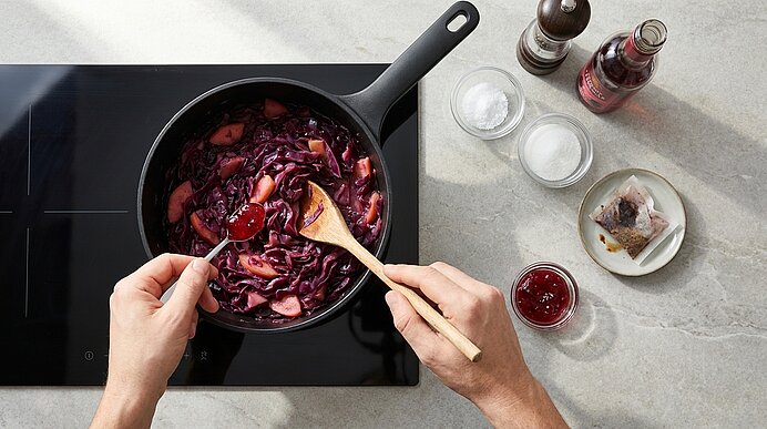 Zwei Hände rühren in einer Pfanne auf dem Herd geschmorten Rotkohl mit einem Holzlöffel; daneben kleine Schälchen mit Gewürzen und Zutaten.