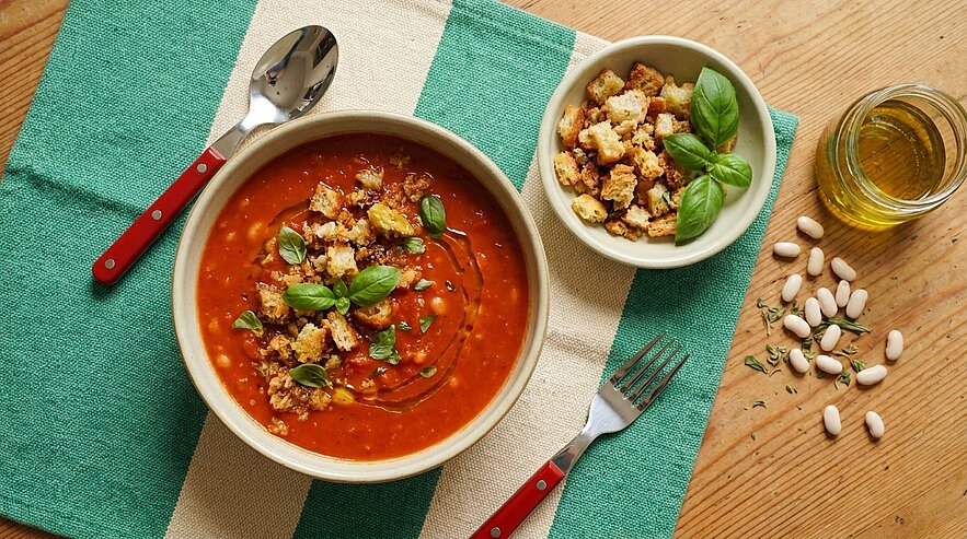 Rote Tomatensuppe in einer Schüssel, garniert mit Basilikumblättern, Croutons und Olivenöl; daneben ein kleines Schälchen mit zusätzlichen Croutons.