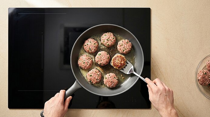 Hände wenden Frikadellen mit Pfannenwender in einer Pfanne auf einem Kochfeld; daneben Schüssel mit rohen Hackfleischbällchen.
