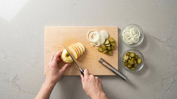 Zwei Hände schneiden einen Apfel in Scheiben auf einem Holzbrett; daneben liegen Zwiebelringe, Cornichons in Schälchen sowie ein Messer und ein Schäler.