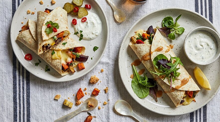 Zwei Teller mit gefüllten Tortilla-Wraps (geröstetes Gemüse, geschmolzener Käse, Blattsalat), dazu ein Schälchen Kräuter-Joghurt-Dip und eine Zitronenscheibe.
