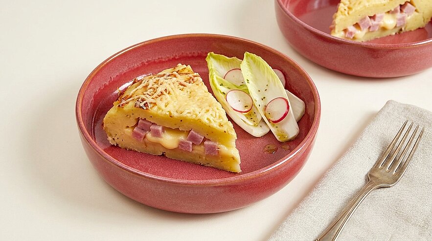 Dreieckiges Stück einer herzhaften Quiche mit geschmolzenem Käse und Schinkenwürfeln auf einem roten Teller, serviert mit Chicoréeblättern und Radieschenscheiben.