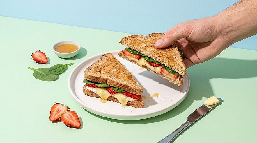 Hand nimmt eine Sandwichhälfte eines Vollkorn-Sandwichs mit Erdbeerscheiben, Blattspinat und geschmolzenem Käse auf weißem Teller.
