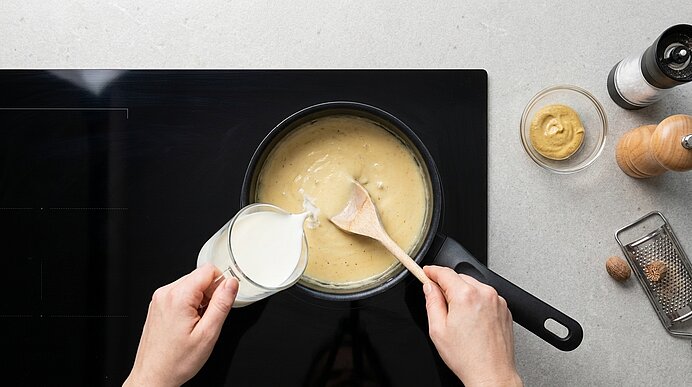 Zwei Hände gießen Milch in eine cremige Sauce im Stieltopf auf dem Kochfeld und rühren mit einem Kochlöffel.