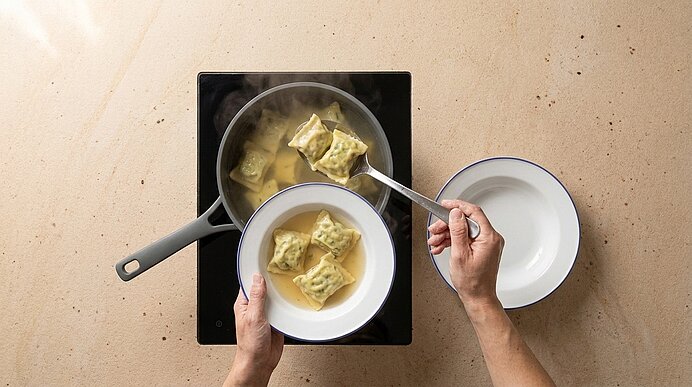 Von oben gesehen: Hände schöpfen dampfende, gefüllte Teigtaschen (Ravioli) mit einer Schaumkelle aus einem Topf Brühe in eine Servierschüssel.