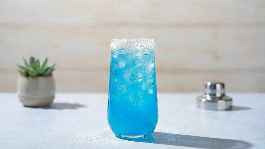 Ein Glas mit blauer, eisgekühlter Flüssigkeit steht auf einem Tisch. Der Rand des Glases ist mit weißem Zucker bedeckt. Im Hintergrund ist ein kleiner Topf mit einer Pflanze zu sehen.