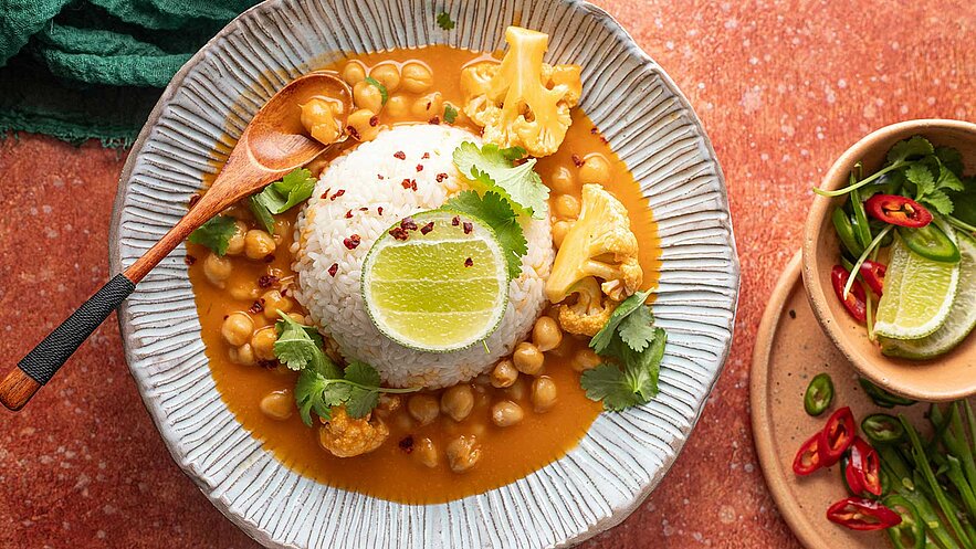 Ein Teller mit Basmati-Reis, umgeben von Kichererbsen und Blumenkohl in einer orangefarbenen Sauce. Frische Kräuter und eine Limettenscheibe dekorieren das Gericht, das mit roten Chiliflocken bestreut ist. Der Teller hat eine leicht gewellte Form und steht auf einem rustikalen Untergrund.