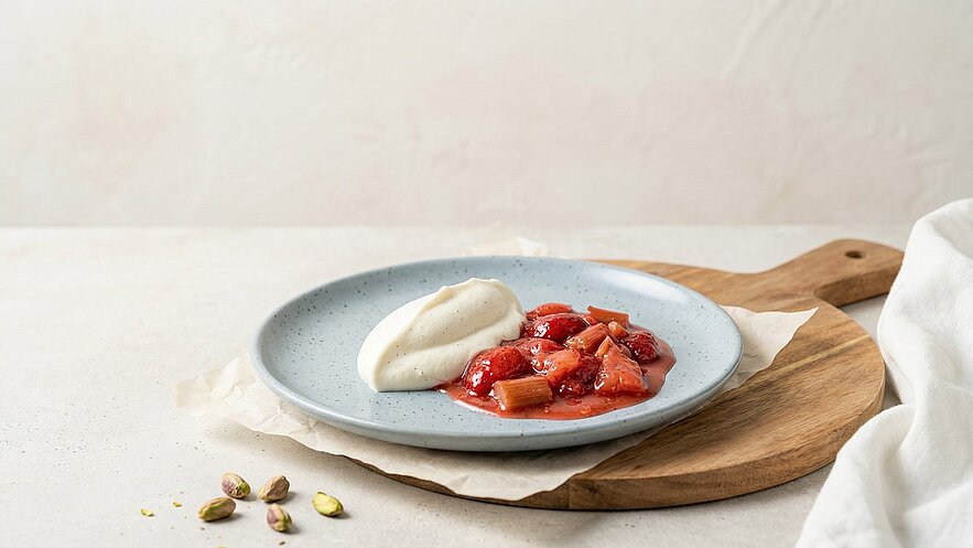Ein grauer Teller auf einem Holzbrett, gefüllt mit Erdbeeren und Rhabarber in rotem Sirup, dazu ein Klecks Vanillecreme. Daneben liegen einige Pistazienkerne. Der Hintergrund ist leicht verschwommen.