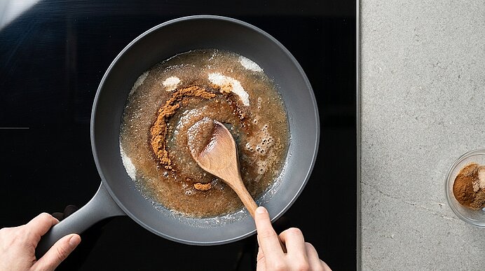 Aufsicht: Zwei Hände rühren mit einem Holzlöffel in einer Pfanne auf dem Herd eine schmelzende braune Zucker‑Gewürz‑Mischung.