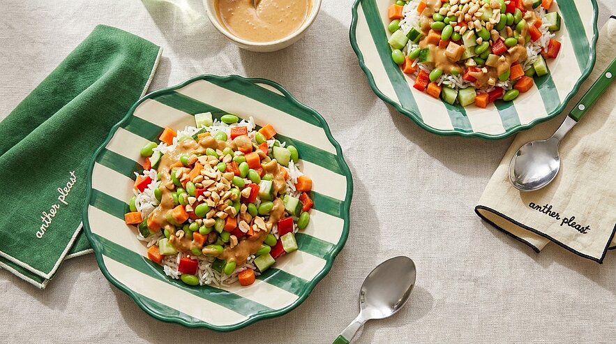 Zwei grün-weiß gestreifte Teller mit buntem Gemüse- und Reisgericht: gewürfelte Gurke, Tomate und Karotte, Edamame, Mais, Erdnüsse und cremiges Dressing; Besteck und Servietten am Tisch.
