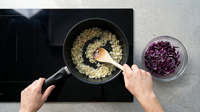 Hände rühren in einer Pfanne auf dem Herd angebratene Zwiebelwürfel; daneben eine Schüssel mit gehobeltem Rotkohl.