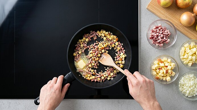 Zwei Hände braten Zwiebel-, Schinken- und Apfelwürfel mit Butter in einer Pfanne auf dem schwarzen Kochfeld an.