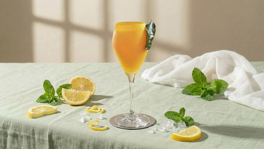 Ein Glas mit einem orangefarbenen Getränk steht auf einem hellgrünen Tisch. Um das Glas sind frische Minzblätter, Zitronenscheiben und einige Eiswürfel verteilt. Im Hintergrund ist ein heller, neutraler Raum sichtbar.