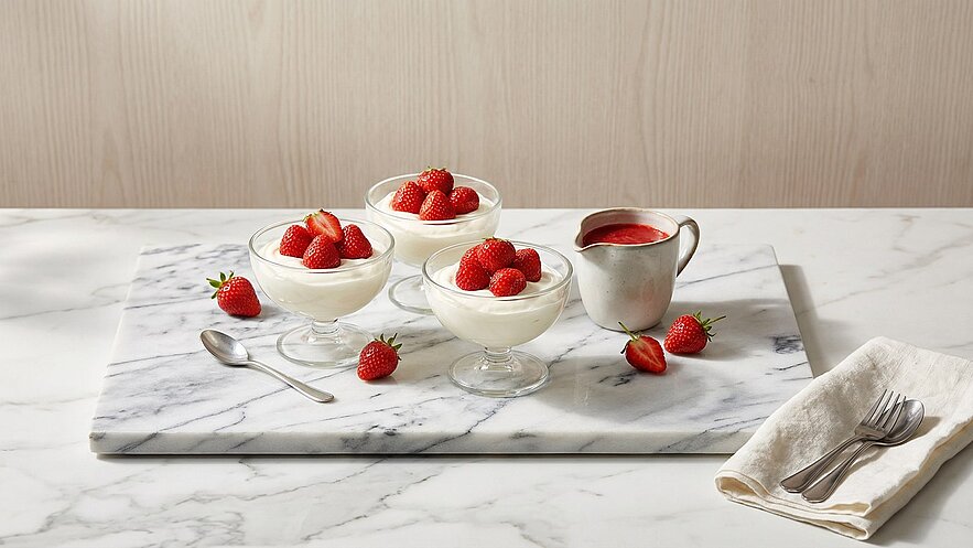 Drei gläserne Schalen mit cremigem Dessert, garniert mit frischen Erdbeeren, stehen auf einem Marmorbrett. Neben den Schalen befindet sich eine kleine Kanne mit Erdbeersauce und einige Erdbeeren sind verstreut. Ein Besteckset liegt bereit.