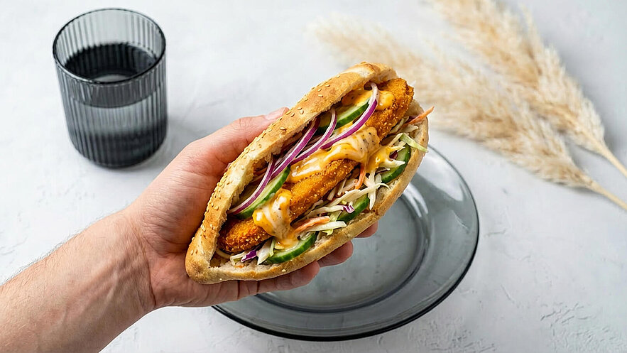 Ein Hand hält ein Sandwich mit paniertem Hühnchen, frischem Gemüse und Sauce. Das Sandwich ist in einem Brötchen serviert. Im Hintergrund steht ein Glas Wasser auf einem Teller. Die Gesamtkomposition ist modern und ansprechend.