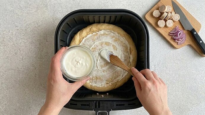 Zwei Hände bestreichen den Brezelboden in der Heißluftfritteuse mit Schmand. Rechts liegen Zwiebel- und Weißwurstscheiben.