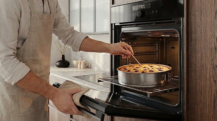 Person in Schürze nimmt einen Obstkuchen mit Ofenhandschuh aus einem Backofen und prüft ihn mit einem Holzspieß.