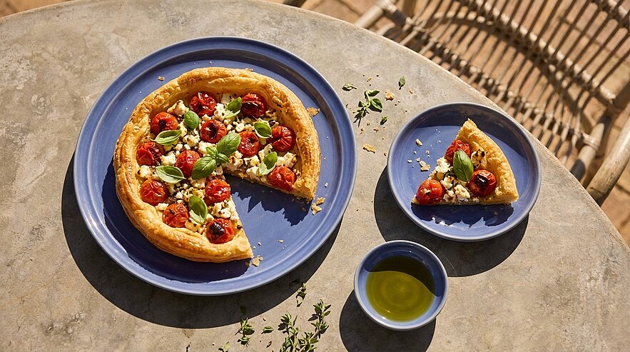 Blätterteigtarte mit gerösteten Kirschtomaten, Feta und Basilikum auf blauem Teller; ein Stück auf kleinem Teller daneben und eine Schale Olivenöl, auf hellem Tisch serviert.