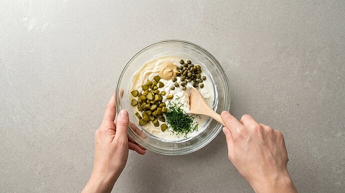 Zwei Hände rühren in einer Glasschüssel cremige Sauce mit gehackten Cornichons, Kapern und frischem Dill.