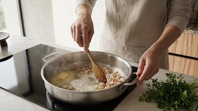 Hände einer Person in Schürze rühren mit einem Holzlöffel kleine Knoblauchstücke in einer Pfanne mit schmelzender Butter auf dem Herd.