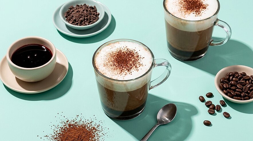 Zwei Glasbecher Cappuccino mit Milchschaum und Kakaopulver auf türkisfarbenem Untergrund, umgeben von einer kleinen Tasse mit schwarzem Kaffee, Schalen mit Kaffeebohnen und Schokoladenraspeln, einem Löffel und verstreutem Kaffeepulver.