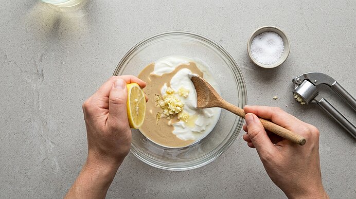 Zwei Hände bereiten eine Sauce: in einer Glasschüssel werden Joghurt und Tahini mit gehacktem Knoblauch vermischt, eine Hand presst Zitrone hinein.