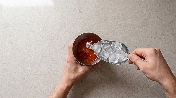 Zwei Hände schaufeln Eiswürfel mit einer Metallschaufel in ein Glas mit braunem Cocktail.