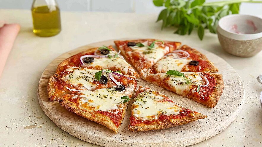 Eine frisch gebackene Pizza auf einem runden Holzbrett. Sie ist belegt mit Tomatensoße, geschmolzenem Käse, schwarzen Oliven und frischen Kräutern. Einige Stücke sind bereits herausgeschnitten und angerichtet.